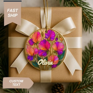 May include: A round Christmas ornament with a floral design in shades of pink, purple, and orange, with the name "Olivia" in a script font. The ornament hangs from a gold string and is set against a wrapped gift with a cream ribbon.