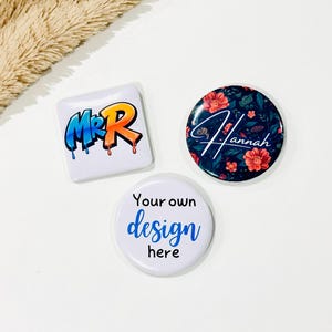 May include: Three custom pin-back badges. One square badge features graffiti-style text reading Mr R. One circular badge has a dark floral pattern with the name Hannah in white script. One circular badge is white with the text Your own design here.