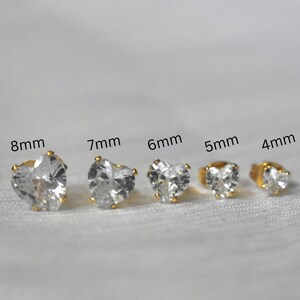 May include: A collection of heart-shaped stud earrings in various sizes, ranging from 4mm to 8mm. Each earring features a clear, faceted stone set in a gold-colored metal. The earrings are displayed on a white, textured surface.