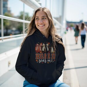 Op de afbeelding: Een zwarte hoodie met een grafische print van drie pompoenen versierd met strikken met patronen. De tekst "HOARD season" staat onder de pompoenen in een decoratief lettertype, omringd door kleine oranje en bruine herfstbladeren.