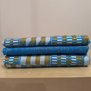 May include: Three folded layers of woven fabric featuring a vibrant blue base colour. The textile displays a repeating geometric pattern composed of alternating white and metallic gold rectangular blocks integrated into the blue weave throughout.