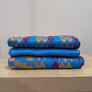 May include: A stack of three folded pieces of blue fabric. The top and bottom pieces feature a repeating geometric pattern in shades of yellow, pink, and dark red. The middle piece is a solid, textured blue fabric with a subtle sheen.