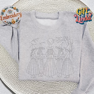 May include: A light gray sweatshirt featuring a line drawing of four figures in witch hats and long skirts, holding a banner that reads "Girls Will Be Girls." The sweatshirt is displayed on a decorative plate.