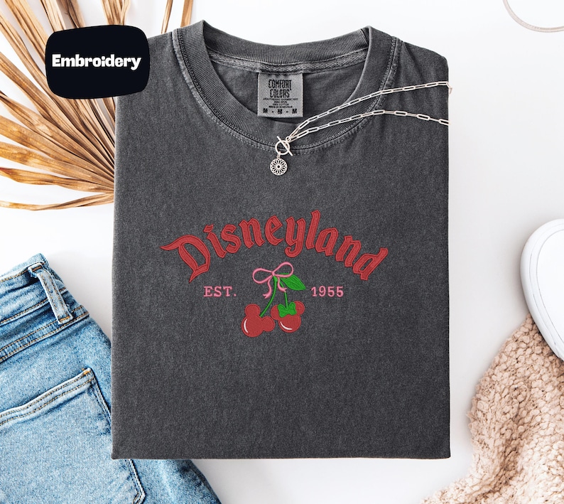 a gray t-shirt with a Disneyland logo, a pair of blue jeans, and some accessories such as a necklace and a scarf, all laid out on a white surface.