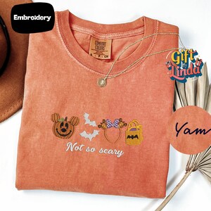 a red t-shirt with a Halloween-themed embroidered design, including a pumpkin, bats, and a cat. The text on the t-shirt says "Not so scary".