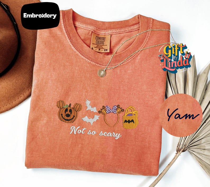 a red t-shirt with a Halloween-themed embroidered design, including a pumpkin, bats, and a cat. The text on the t-shirt says "Not so scary".