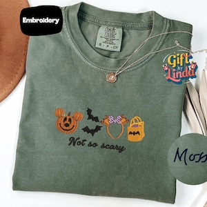 a green t-shirt with a Halloween-themed embroidered design, including a pumpkin, bats, and a bag. The text on the t-shirt says "Not so scary".