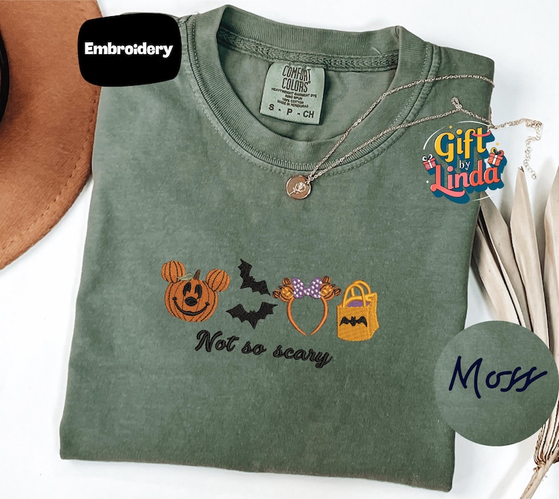 a green t-shirt with a Halloween-themed embroidered design, including a pumpkin, bats, and a bag. The text on the t-shirt says "Not so scary".