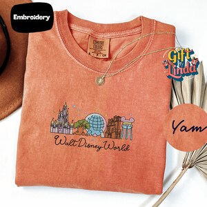 a red t-shirt with a graphic design of Walt Disney World, including a castle, globe, and other iconic landmarks. The t-shirt is displayed on a white background, with a small tag attached to it. There is also a small circular logo with the text "Yam" written on it, and a small brown hat is visible in the background.