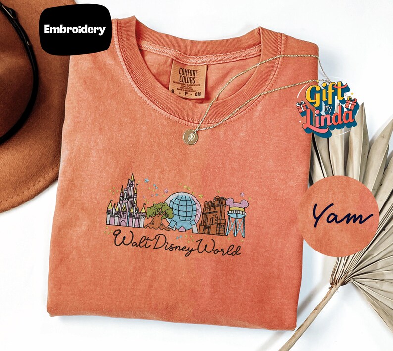 a red t-shirt with a graphic design of Walt Disney World, including a castle, globe, and other iconic landmarks. The t-shirt is displayed on a white background, with a small tag attached to it. There is also a small circular logo with the text "Yam" written on it, and a small brown hat is visible in the background.