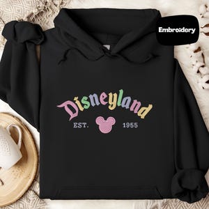 A black hoodie with the word "Disneyland" and the year "1955" printed on it, along with a pink Mickey Mouse logo. The hoodie is placed on a white surface, and there is a mug and some white flowers in the background.