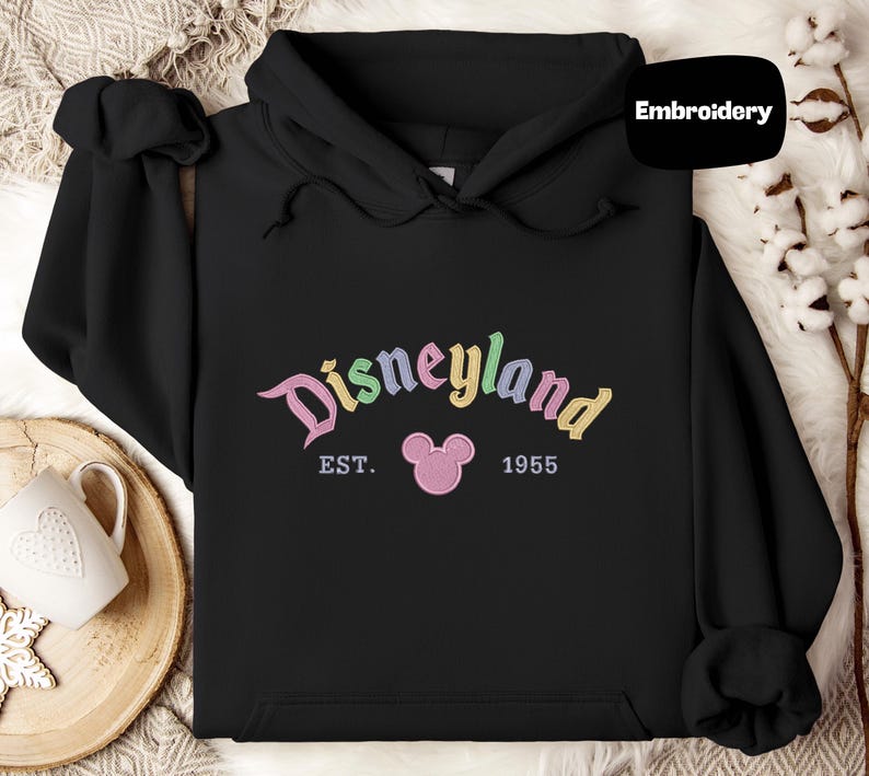 A black hoodie with the word "Disneyland" and the year "1955" printed on it, along with a pink Mickey Mouse logo. The hoodie is placed on a white surface, and there is a mug and some white flowers in the background.