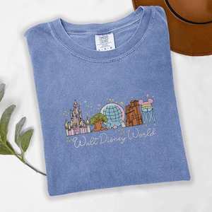 A blue t-shirt with a graphic design of various Disney landmarks, including the Eiffel Tower, Walt Disney World, and other iconic structures, is displayed on a white surface alongside a brown hat.