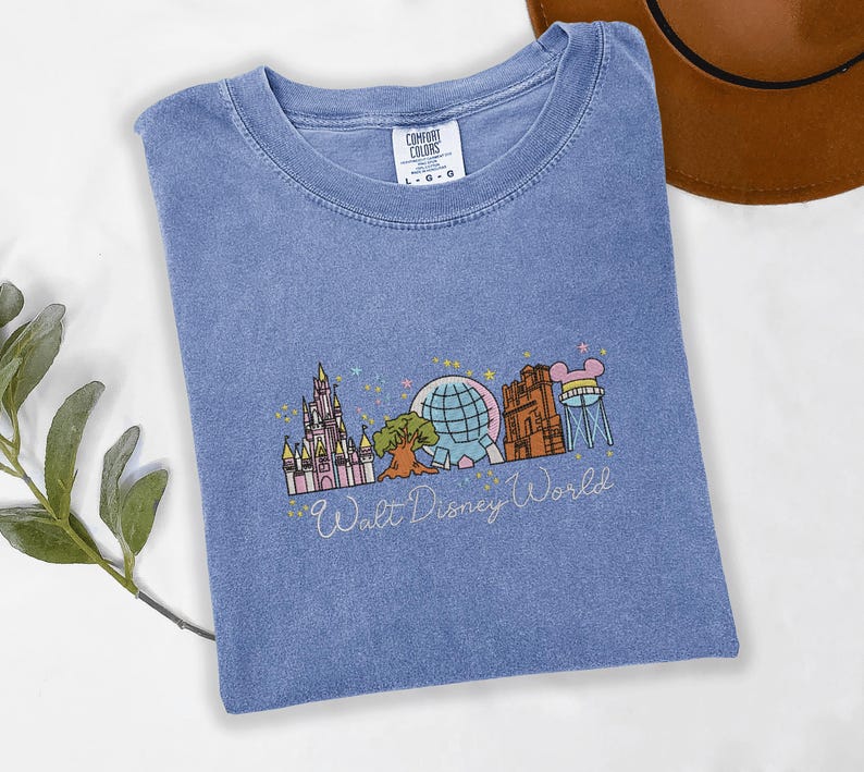 A blue t-shirt with a graphic design of various Disney landmarks, including the Eiffel Tower, Walt Disney World, and other iconic structures, is displayed on a white surface alongside a brown hat.