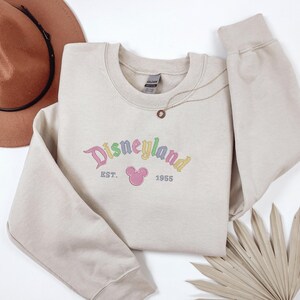 A beige sweatshirt with the text "Disneyland EST. 1955" printed on it, along with a Mickey Mouse logo, is displayed on a white background. A brown hat and a fan are also present in the image.