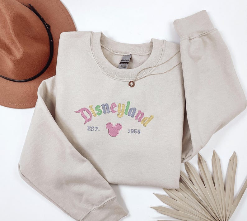 A beige sweatshirt with the text "Disneyland EST. 1955" printed on it, along with a Mickey Mouse logo, is displayed on a white background. A brown hat and a fan are also present in the image.