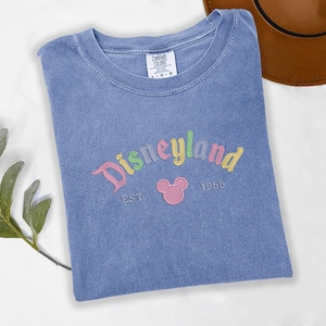 A blue t-shirt with the text "Disneyland EST 1956" printed on it, along with a Mickey Mouse logo, is displayed on a white surface.