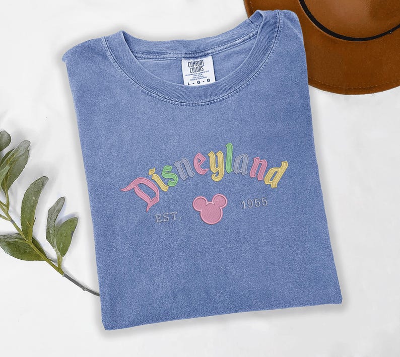 A blue t-shirt with the text "Disneyland EST 1956" printed on it, along with a Mickey Mouse logo, is displayed on a white surface.