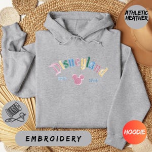 a gray hoodie with a Disneyland logo, a hat, and some straw items, all arranged on a woven mat.