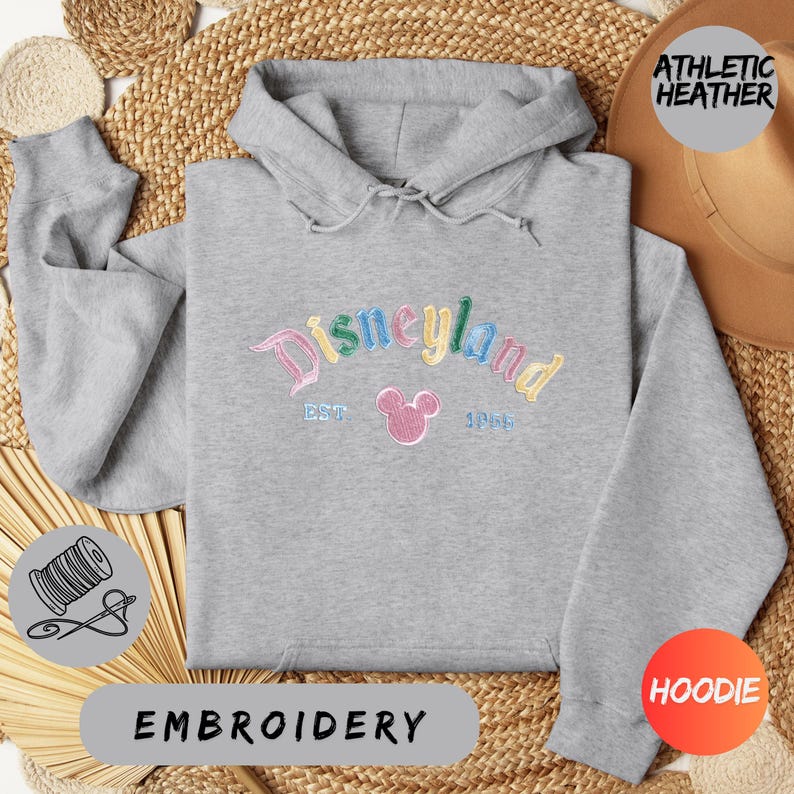 a gray hoodie with a Disneyland logo, a hat, and some straw items, all arranged on a woven mat.