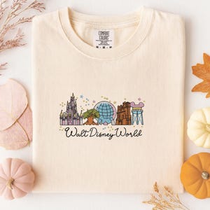 A white t-shirt with a graphic design featuring various Disney landmarks and the text "Walt Disney World" printed on it, surrounded by autumn leaves and small pumpkins.
