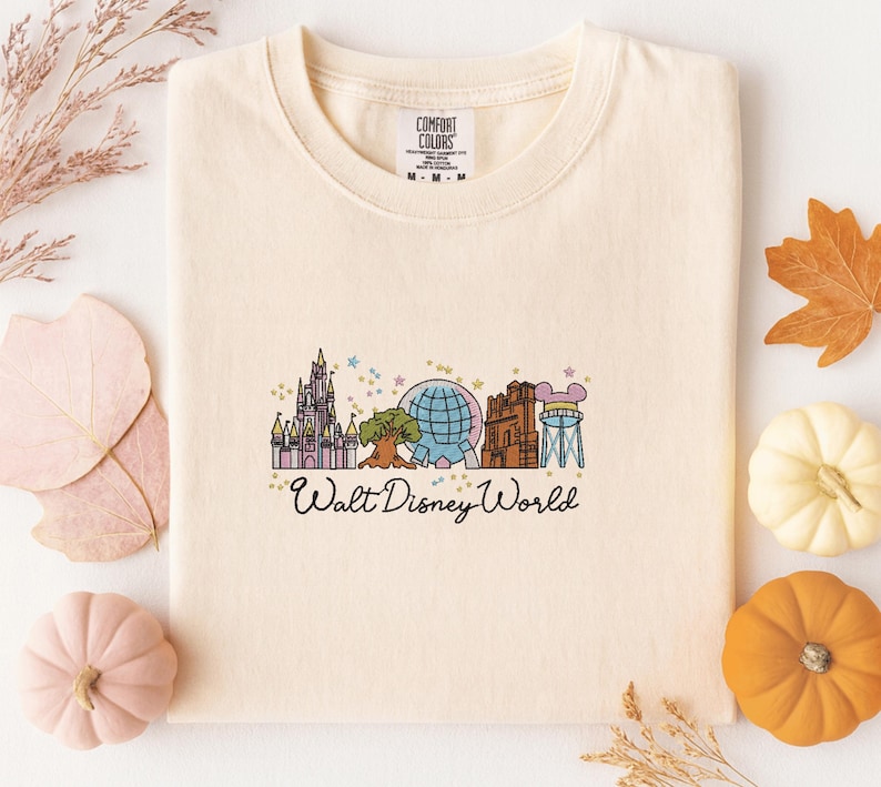 A white t-shirt with a graphic design featuring various Disney landmarks and the text "Walt Disney World" printed on it, surrounded by autumn leaves and small pumpkins.