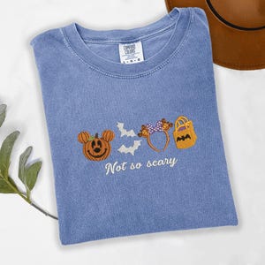 A blue t-shirt with a Halloween-themed design, including a pumpkin, bats, and a bag, along with the text "Not so scary" printed on it.