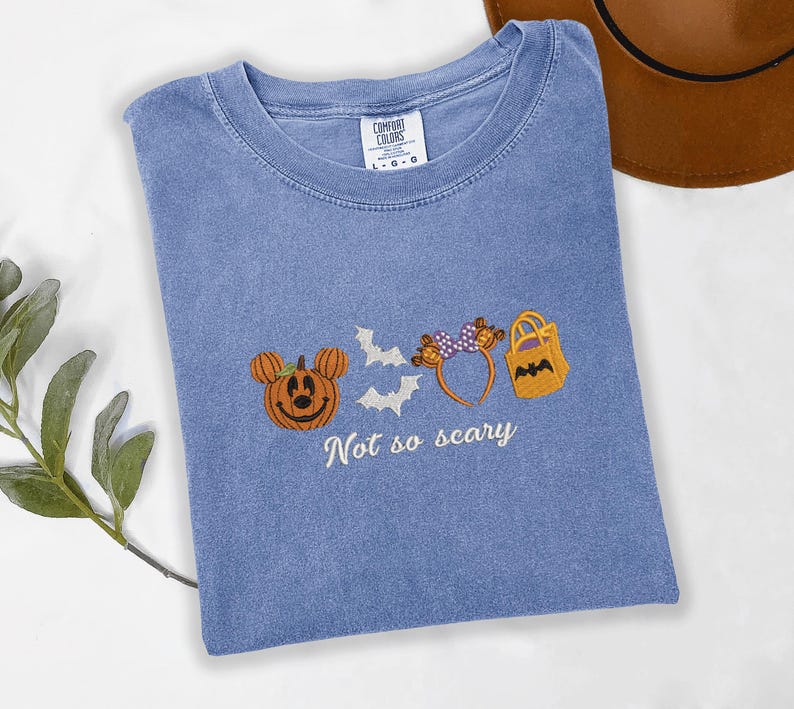 A blue t-shirt with a Halloween-themed design, including a pumpkin, bats, and a bag, along with the text "Not so scary" printed on it.