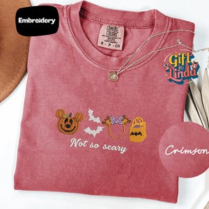 a pink t-shirt with a Halloween-themed embroidered design, including a pumpkin, bats, and a bat-shaped bag. The text on the t-shirt says "Not so scary" and is accompanied by a small circular logo with the text "Crimson".