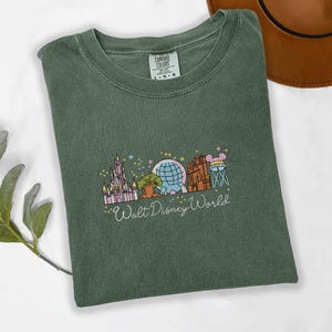 A green t-shirt with a graphic design of various Disney landmarks, including the Eiffel Tower, Walt Disney World, and other iconic structures, is displayed on a white surface alongside a brown hat.