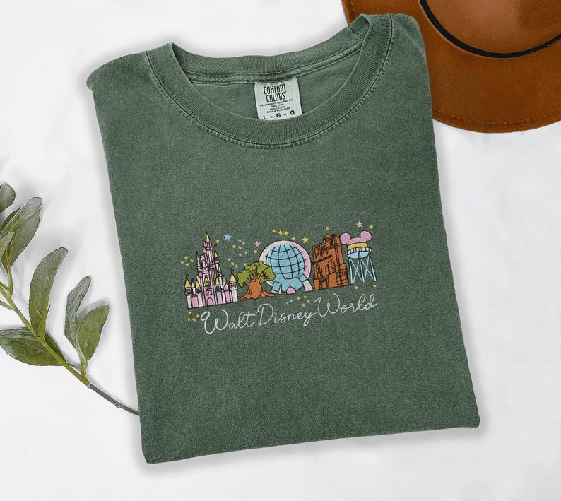 A green t-shirt with a graphic design of various Disney landmarks, including the Eiffel Tower, Walt Disney World, and other iconic structures, is displayed on a white surface alongside a brown hat.