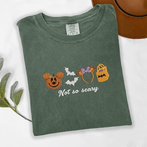 A green t-shirt with a Halloween-themed design, including a pumpkin, bats, and a bag, along with the text "Not so scary" printed on it.