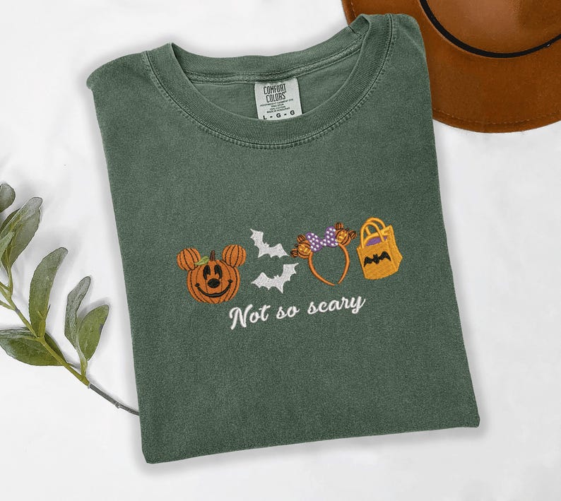 A green t-shirt with a Halloween-themed design, including a pumpkin, bats, and a bag, along with the text "Not so scary" printed on it.