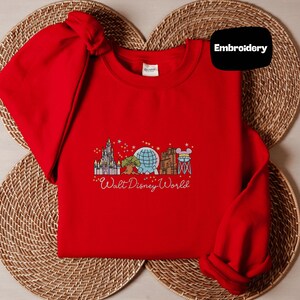 A red t-shirt with a graphic design of Disney landmarks and the text "Walt Disney World" printed on it, placed on a woven placemat.