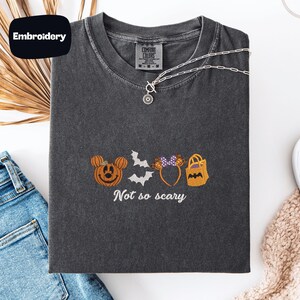 a gray t-shirt with a Halloween-themed embroidered design, including a pumpkin, bats, and a bag. The text on the t-shirt says "Not so scary".
