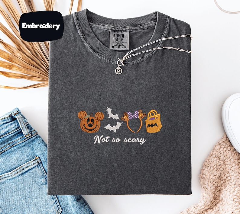 a gray t-shirt with a Halloween-themed embroidered design, including a pumpkin, bats, and a bag. The text on the t-shirt says "Not so scary".