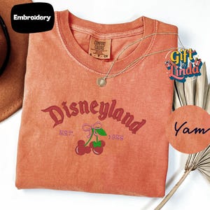a red t-shirt with the text "Disneyland EST. 1955" and cherries printed on it, along with a necklace and a fan.