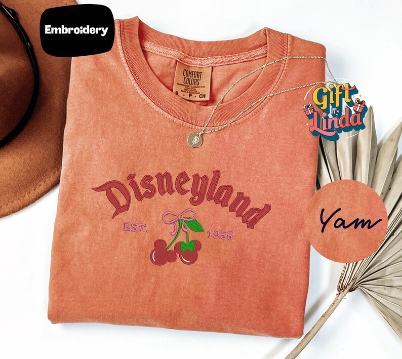 a red t-shirt with the text "Disneyland EST. 1955" and cherries printed on it, along with a necklace and a fan.