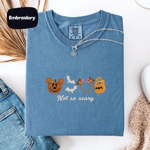 a blue t-shirt with a Halloween-themed embroidered design, including a pumpkin, bats, and a bat-shaped bag. The phrase "Not so scary" is also embroidered on the t-shirt.