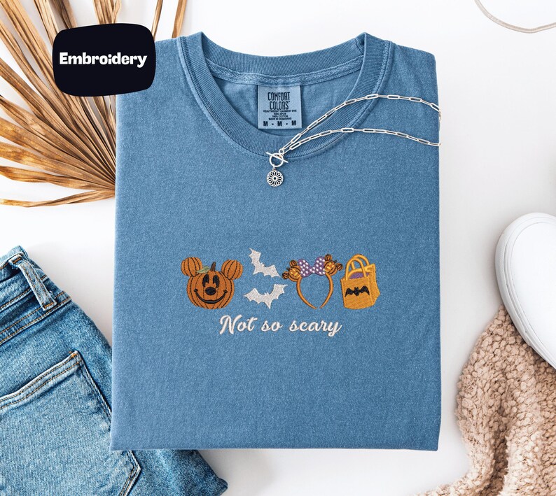 a blue t-shirt with a Halloween-themed embroidered design, including a pumpkin, bats, and a bat-shaped bag. The phrase "Not so scary" is also embroidered on the t-shirt.