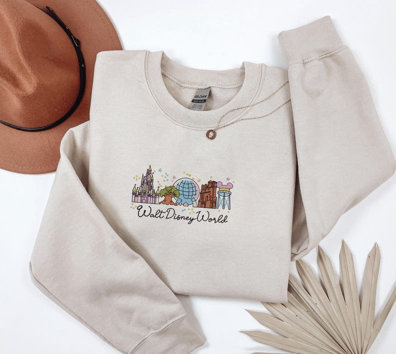 A beige sweatshirt with a graphic of Walt Disney World landmarks, a brown hat, and a fan are arranged on a white background.