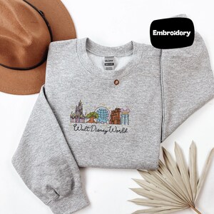 a gray sweatshirt with a Disney World-themed embroidered design, a brown hat, and a fan, all laid out on a white background.