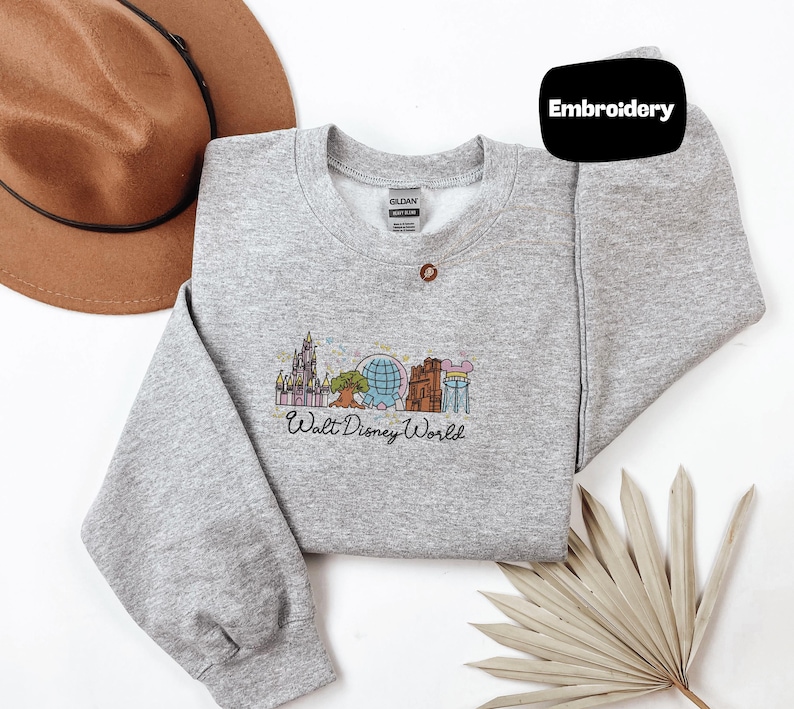 a gray sweatshirt with a Disney World-themed embroidered design, a brown hat, and a fan, all laid out on a white background.