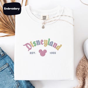 a white t-shirt with a Disneyland logo, a pair of blue jeans, and some decorative elements such as a necklace and a beige scarf.