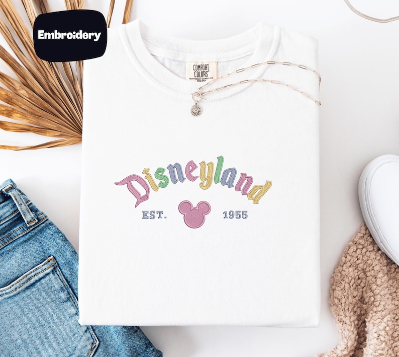 a white t-shirt with a Disneyland logo, a pair of blue jeans, and some decorative elements such as a necklace and a beige scarf.