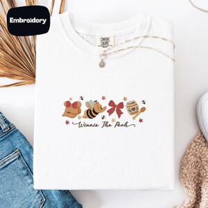 May include: White t-shirt with an embroidered Winnie the Pooh design. The design includes a bow, a bee, a red bow, a honey pot, and the text "Winnie The Pooh". The shirt has a round neckline and short sleeves.
