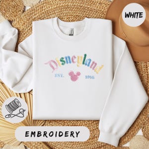 A white sweater with the text "Disneyland EST. 1955" embroidered on it, along with a Mickey Mouse logo, is displayed on a woven straw mat.