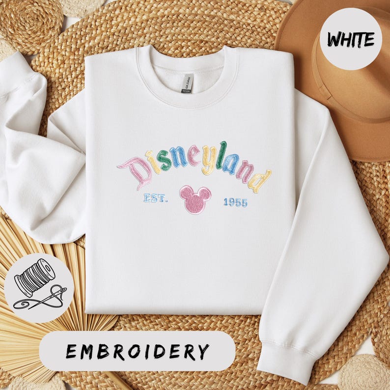 A white sweater with the text "Disneyland EST. 1955" embroidered on it, along with a Mickey Mouse logo, is displayed on a woven straw mat.