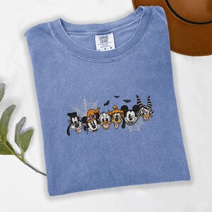 May include: A blue short-sleeve t-shirt with a crew neckline and a Comfort Colors label. The centre features an embroidered graphic of various cartoon characters wearing Halloween costumes, including spider webs and small black bats above them.
