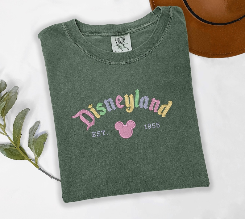 A green t-shirt with the text "Disneyland EST. 1955" printed on it, along with a Mickey Mouse logo, is displayed on a white surface.